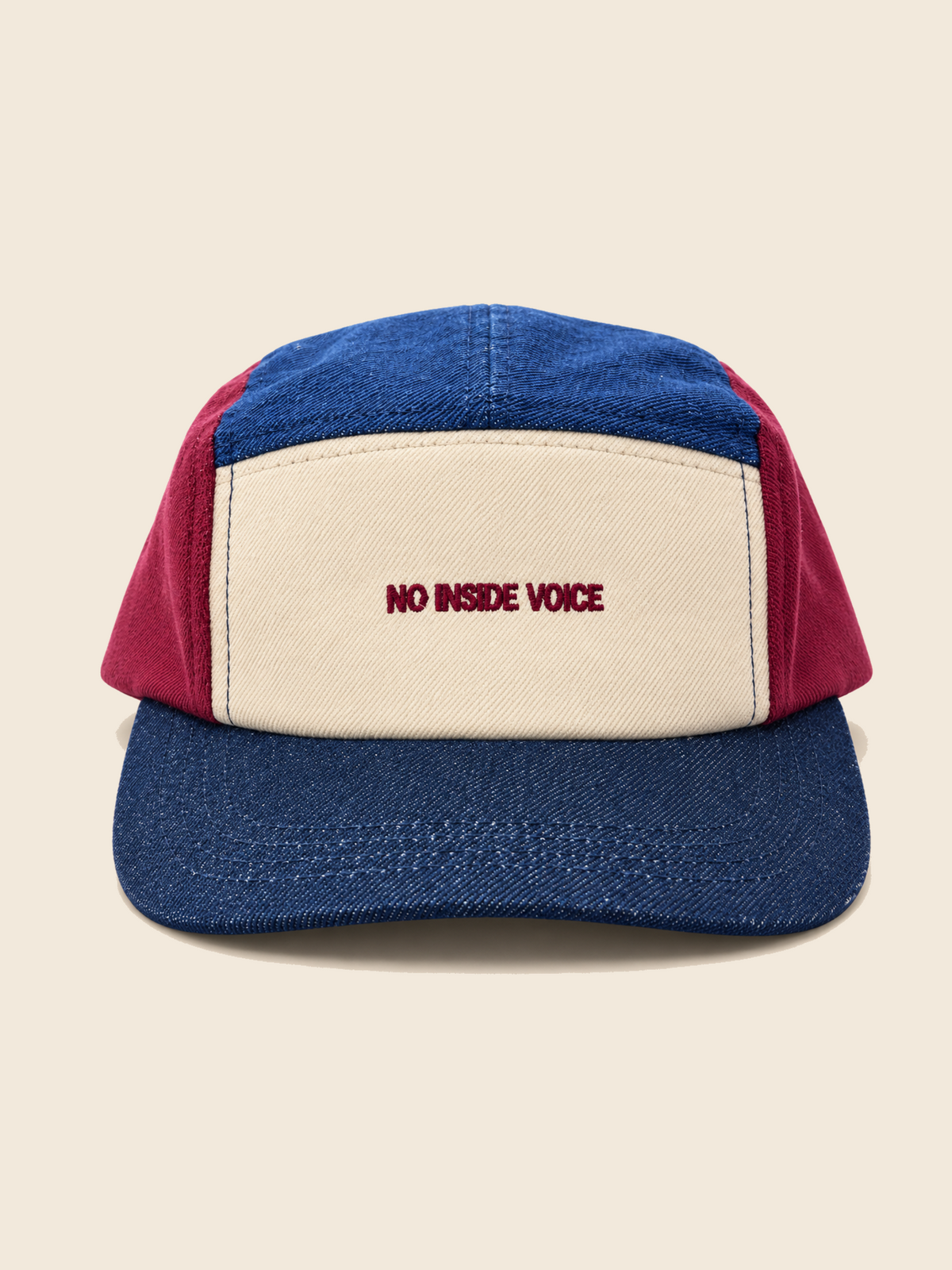 A colorblock 5-panel  kids hat with a blue denim-like front panel and brim, a cream canvas front panel, and red side panels. The front panel features the embroidered text 'NO INSIDE VOICE' in red.