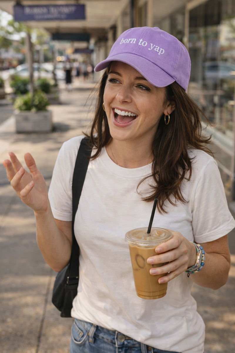 Woman wearing a purple cap with 'Born to yap' text, holding a drink, and walking outdoors.